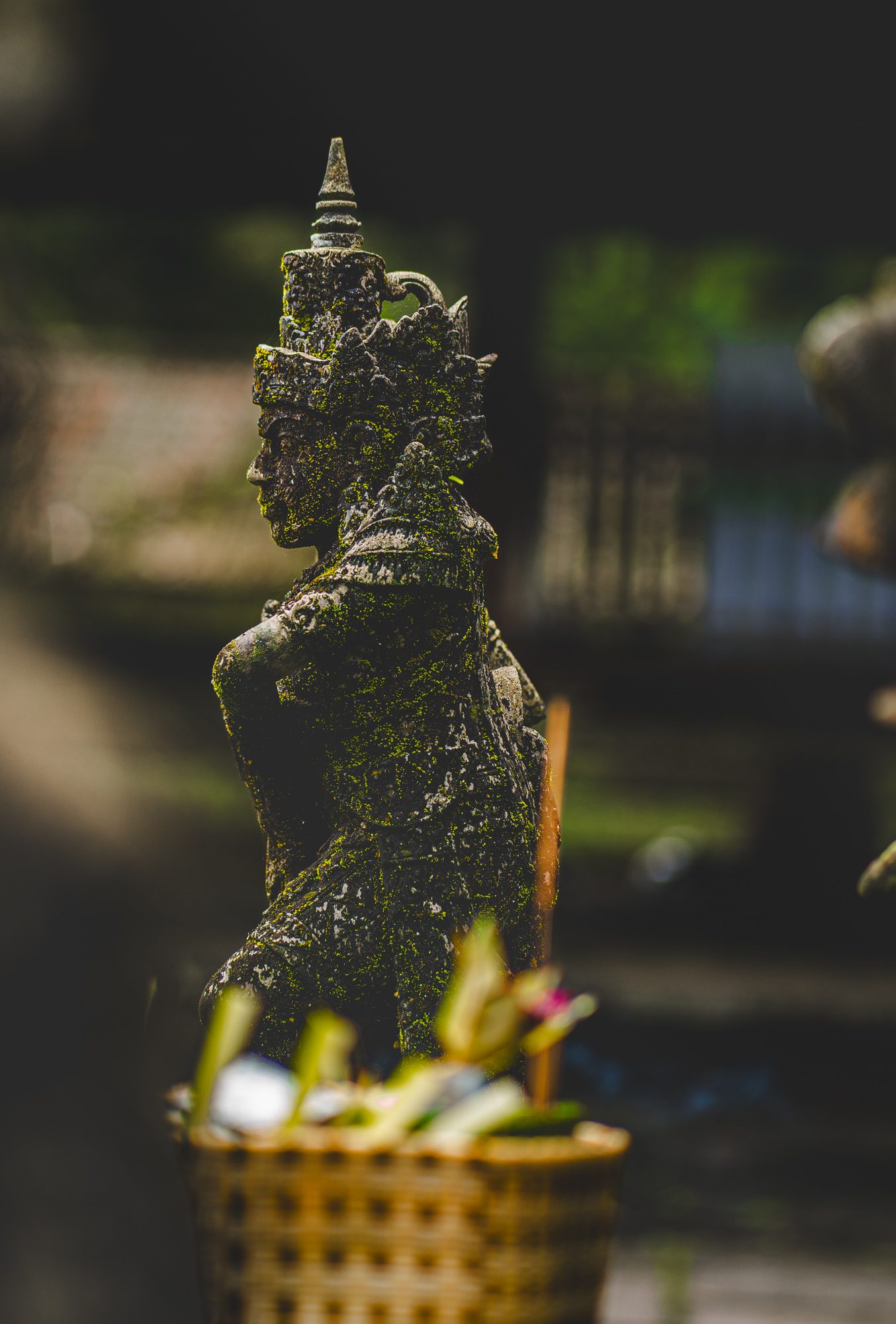 Statue en pierre couverte de mousse dans un temple balinais, offrandes de fleurs au premier plan